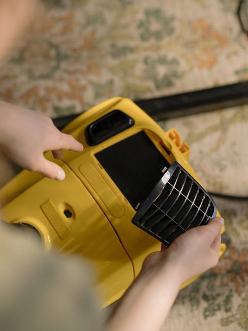 A person holding and preparing to operate a yellow industrial carpet cleaning machine with a black handle and vent, positioned over a patterned area rug with muted tones of beige, green, and orange, i