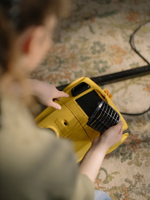 A person holding and preparing to operate a yellow industrial carpet cleaning machine with a black handle and vent, positioned over a patterned area rug with muted tones of beige, green, and orange, i