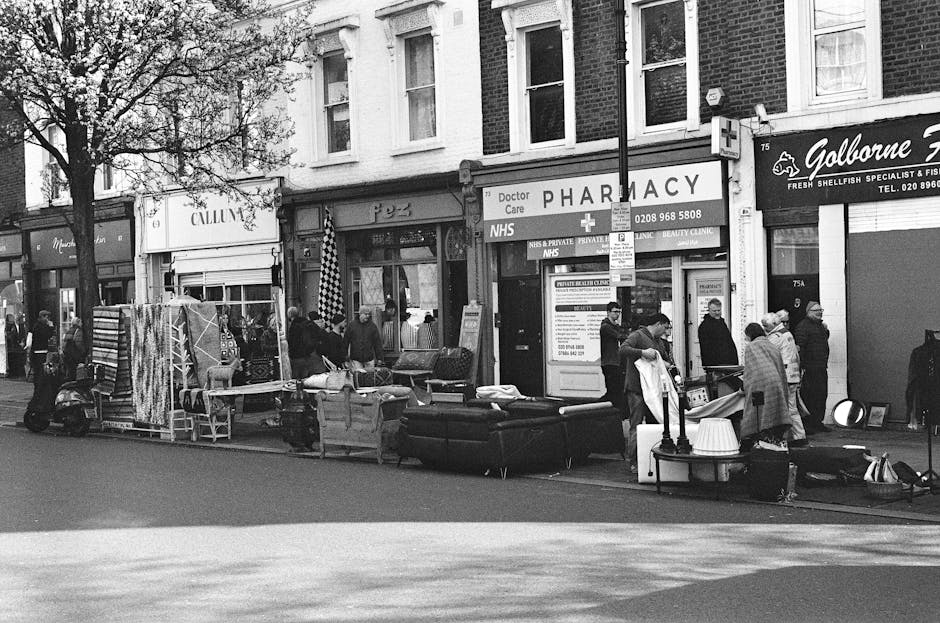 A black-and-white street scene outside shops and a pharmacy, featuring multiple people browsing and shopping. Furniture and household items, including sofas, chairs, and a table with a lamp, are displ
